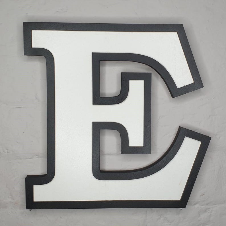 May include: A large, white, uppercase letter "E" with a black outline. The letter is made of wood and has a slightly distressed finish.