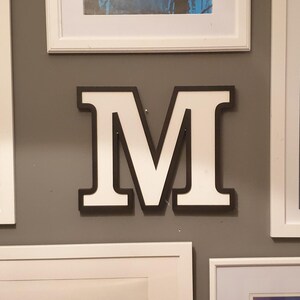 May include: A large white letter M with a black outline is mounted on a gray wall. The letter is surrounded by white picture frames.