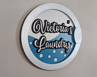 May include: A round, silver-framed sign with a blue and white interior. The sign says "Victoria's Laundry" in black and white lettering.