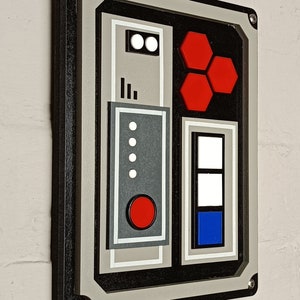 May include: A rectangular wall art piece with a black frame and a grey, black, and white design. The design includes red hexagons, white circles, and a red circle. The art is mounted on a white brick wall.