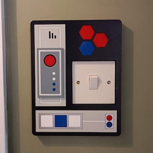 May include: A black wall plate with a retro-futuristic design. It features a white light switch, red and blue hexagonal shapes, and a panel with a large red circle and smaller blue and white circles. The design is reminiscent of a spaceship control panel.