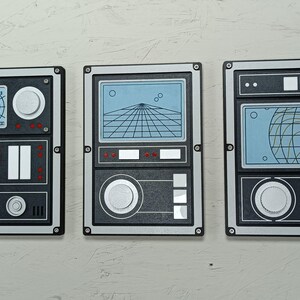 May include: Three retro-style wall art panels with a black, gray, and silver color scheme. Each panel features a different display design, including radar, a grid, and a globe. The panels have a rectangular shape with rounded corners and are mounted on a light-colored wall.