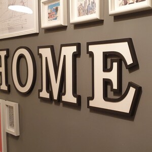 May include: White letters spelling "HOME" with black outlines, mounted on a gray wall. The letters are 3D and have a shadow effect.