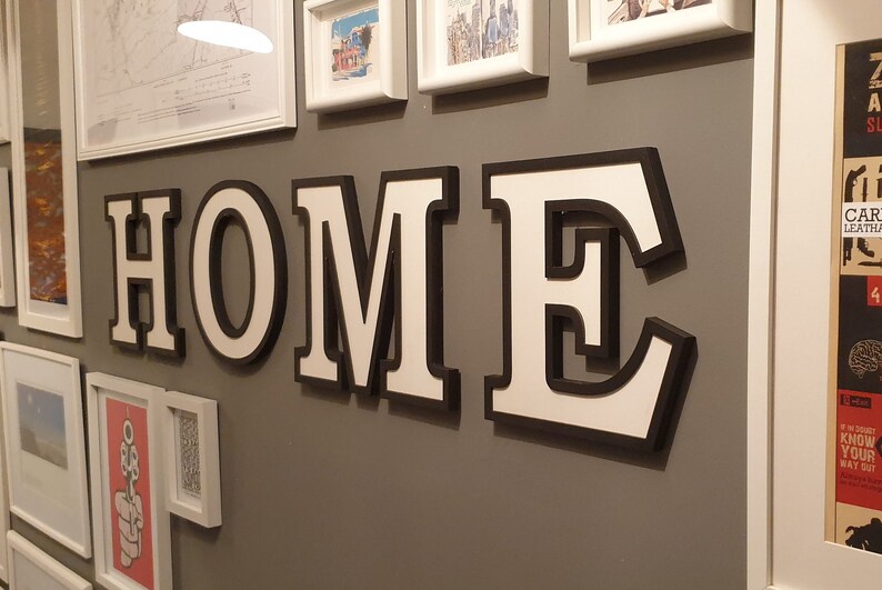May include: White letters spelling "HOME" with black outlines, mounted on a gray wall. The letters are 3D and have a shadow effect.