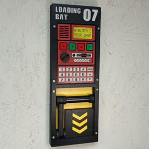 May include: A black and red wall-mounted control panel with the text "LOADING BAY 07" and "AIRLOCK 2 DOOR OPEN." It features a keypad, buttons, and a yellow and black section with chevron arrows. The panel is mounted on a light-colored wall.