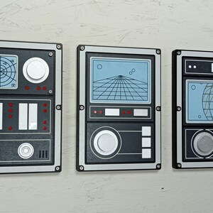May include: Three retro-style wall panels with a black and silver frame. Each panel features a different blue and white graphic design, resembling vintage radar or control panels. The panels are mounted on a light-colored wall.
