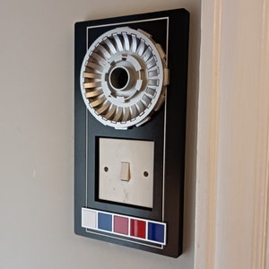May include: A black light switch cover with a silver decorative plate featuring a circular design. The cover has a white light switch and a row of red, white, and blue accents.