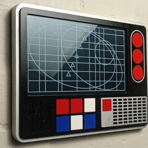 May include: A black and silver wall-mounted radar screen with a grid pattern and triangular markers on the screen. The screen has red, white, and blue buttons and a silver border. The radar screen is mounted on a textured white wall.