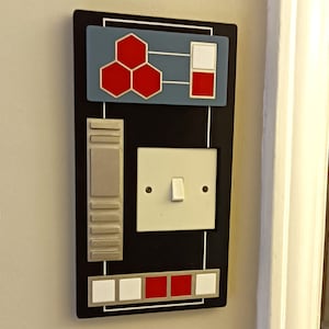 May include: A black light switch cover with a silver panel and a white light switch. The cover has a red, white and blue design with a geometric pattern and the words "Starfleet Command".