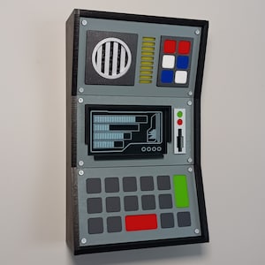 May include: A gray control panel with a black frame, a screen with a digital display, a row of buttons, and a row of colored lights. The panel has a black frame and a gray background. The screen is rectangular and has a black border. The buttons are rectangular and gray. The lights are red, green, and blue.