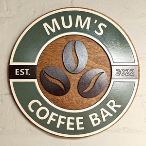 May include: A round wooden sign with a green background and white lettering. The sign says "Mum's Coffee Bar" with the text "EST. 2022" on the top and bottom of the circle. There are three coffee bean shapes in the centre of the sign.