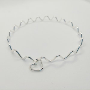 Op de afbeelding: Een zilveren armband met een golvend ontwerp en een klein hartje.