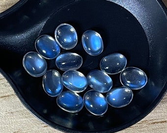 Könnte beinhalten: Eine Sammlung ovaler, durchscheinender blauer Edelsteine mit einem dezenten Glanz, präsentiert in einem schwarzen, schaufelförmigen Behälter. Die Edelsteine haben eine polierte, glatte Oberfläche. Der Behälter steht auf einer hellen Holzoberfläche.