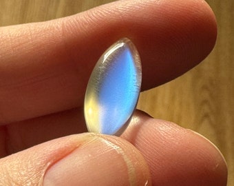 Könnte beinhalten: Ein polierter, oval geformter Edelstein mit schimmerndem Glanz. Der Stein wechselt von einem blassen Gelb zu einem leuchtenden Blau, mit feinen inneren Strukturen. Zwischen zwei Fingern gehalten, reflektiert die Oberfläche des Steins das Licht.