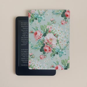 Può includere: Una custodia per e-reader con un motivo floreale di rose rosa, margherite bianche e foglie verdi su sfondo verde chiaro. La custodia è aperta, rivelando lo schermo dell'e-reader. L'e-reader è nero.