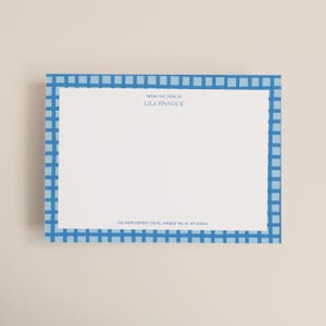 May include: A rectangular stationery card with a blue gingham border. The card features the text "FROM THE DESK OF LILA PINNOCK" and the address "742 MAPLEBERRY DRIVE, AMBER FALLS, WY 62845". The background is white.