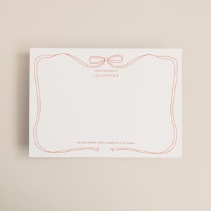 May include: A white stationery card featuring a copper-coloured border and bow design. The text "FROM THE DESK OF LILA PINNOCK" is at the top, with an address at the bottom. The card is rectangular.