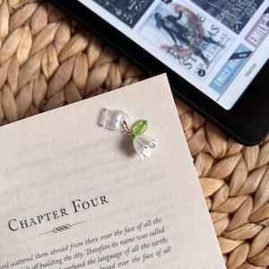 May include: A clear plastic page holder with a gold-coloured charm featuring a white flower and green leaves. The page holder is attached to a book with the text "CHAPTER FOUR" visible. An e-reader is in the background.