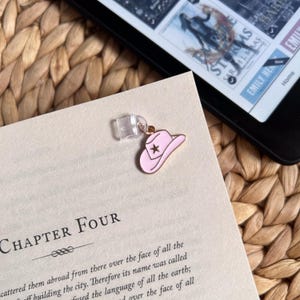 May include: A pink cowboy hat charm with a star detail attached to a clear clip, resting on a book page. The word "CHAPTER FOUR" is visible. An e-reader is in the background.