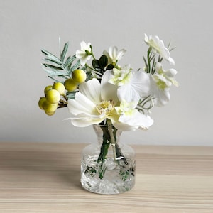 Pode incluir: Um pequeno vaso de vidro transparente com um buquê de flores brancas, folhas verdes e bagas amarelas. As flores têm pétalas grandes e delicadas. O vaso está sobre uma superfície de madeira clara.