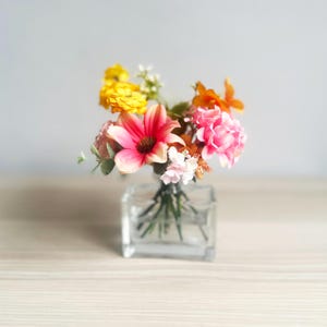 Può includere: Un piccolo vaso quadrato di vetro pieno di una colorata composizione di fiori artificiali. Il bouquet comprende fiori rosa, gialli e arancioni vibranti, con tocchi di bianco e verde. Il vaso è posto su una superficie di legno chiaro.