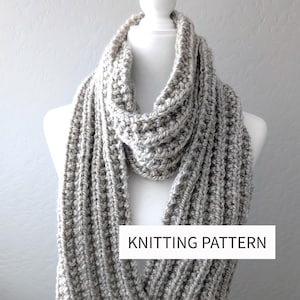 May include: A chunky, light grey, ribbed knit scarf draped on a white mannequin. The scarf features a thick, textured yarn construction with vertical ridges throughout. A white rectangular label with the black text KNITTING PATTERN is visible.