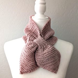 May include: A dusty rose pink knitted scarf featuring a garter stitch texture. The scarf is designed with a pull-through loop mechanism, allowing one end to pass through a slit in the other, creating a secure and layered wrap around the neck.