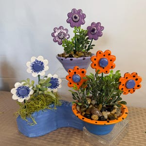 May include: Three ceramic flower arrangements in blue, orange, and purple pots. The flowers have white, orange, and purple petals with blue centers. The pots are filled with greenery and small stones, creating a decorative display.