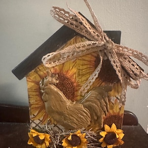 Può includere: Una decorazione in legno a forma di casa con una statuetta di gallo marrone, girasoli e un fiocco di pizzo. La casa è dipinta in uno stile rustico e da fattoria.