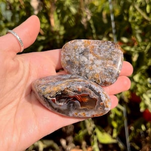 May include: Two polished, smooth, irregular-shaped stones featuring a mottled pattern of gray, white, and earthy brown tones. One stone displays a prominent, hollowed-out center revealing a deep, translucent orange and red crystalline interior.