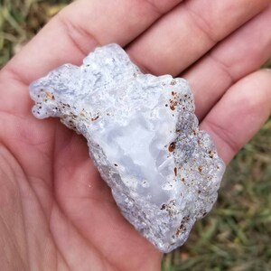 Puede incluir: Una geoda de &aacute;gata blanca y gris con una superficie lisa y pulida. La geoda tiene un exterior natural, rugoso con marcas marrones y grises.