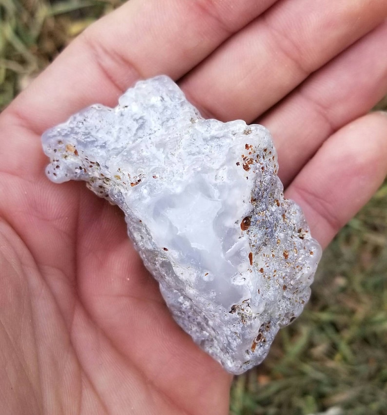 Puede incluir: Una geoda de &aacute;gata blanca y gris con una superficie lisa y pulida. La geoda tiene un exterior natural, rugoso con marcas marrones y grises.
