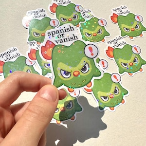Pode incluir: Um conjunto de adesivos verdes e brancos com um design de coruja de desenho animado. A coruja tem uma chama vermelha e laranja atrás e o texto "Spanish or Vanish" no adesivo.