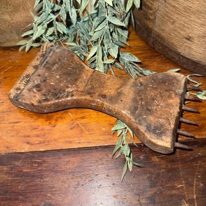 May include: A vintage, wooden tool with a bow-tie shape, featuring a textured surface and metal spikes on each end. The tool is a dark brown color and rests on a wooden surface, with green foliage in the background.