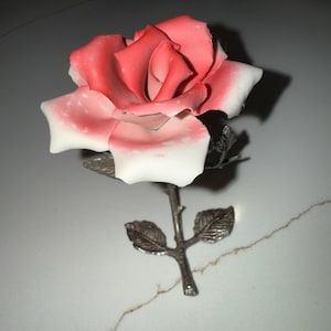 Może przedstawiać: Ceramiczna róża z dwutonową kolorystyką. Płatki przechodzą od żywej czerwieni do miękkiej bieli na krawędziach. Łodyga i liście wykonane są ze srebrnego metalu, tworząc delikatną rzeźbę kwiatową.