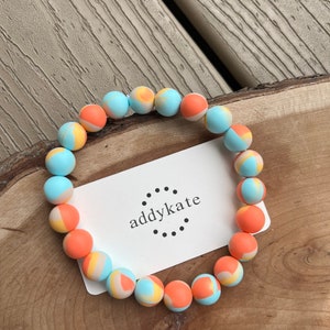 May include: A colorful beaded bracelet with orange, blue, and yellow swirls. The bracelet is on a wooden surface with a white card that says "addykate" in black text.