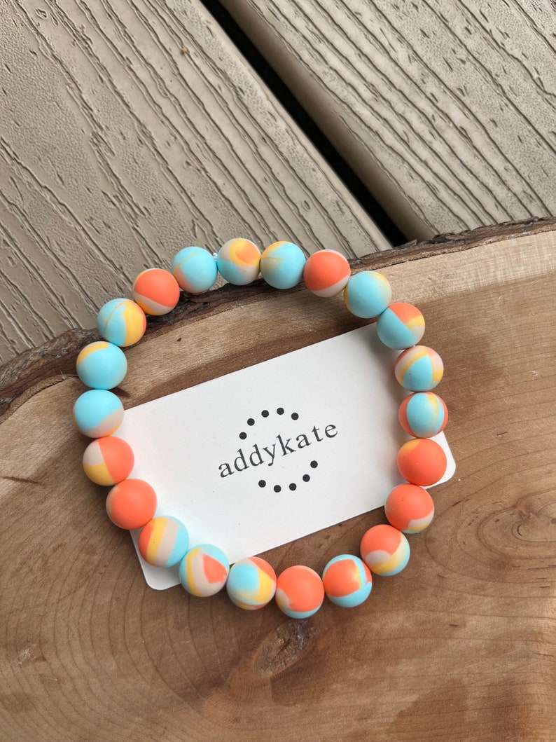 May include: A colorful beaded bracelet with orange, blue, and yellow swirls. The bracelet is on a wooden surface with a white card that says "addykate" in black text.