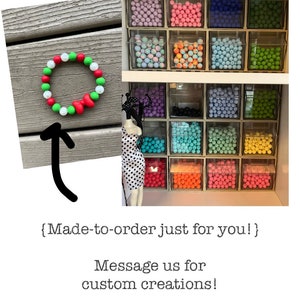 May include: A Christmas wreath bracelet made with green, white, and red silicone beads. The bracelet is displayed on a wooden surface. The image also shows a variety of silicone beads in different colors, organized in clear containers. The text "{Made-to-order just for you!}" and "Message us for custom creations!" is visible below the image.