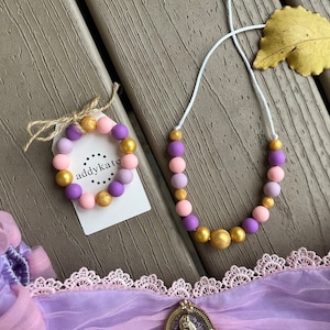 May include: A set of purple, pink, and gold silicone bead jewelry. The set includes a necklace and a bracelet. The necklace is on a white string and the bracelet is tied with a brown ribbon. The bracelet has a small white tag with the text "addykate" on it.
