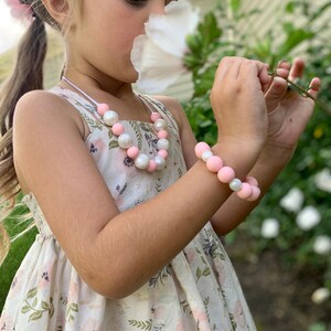 May include: A young girl wearing a white dress with pink and white flowers and a pink and white beaded necklace and bracelet. She is holding a white flower and smelling it.