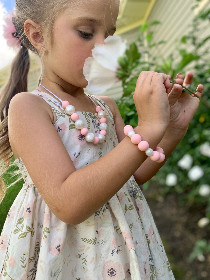 May include: A young girl wearing a white dress with pink and white flowers and a pink and white beaded necklace and bracelet. She is holding a white flower and smelling it.