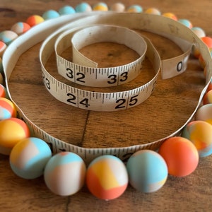May include: A colorful beaded necklace with a mix of blue, orange, and white beads. The necklace is wrapped around a measuring tape that reads 24, 25, 32, and 33 inches.