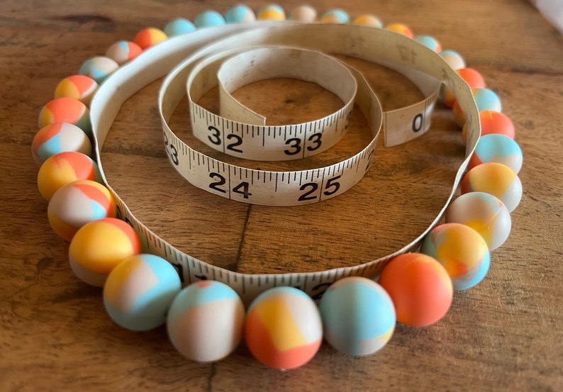 May include: A colorful beaded necklace with a mix of blue, orange, and white beads. The necklace is wrapped around a measuring tape that reads 24, 25, 32, and 33 inches.