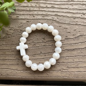 May include: White beaded bracelet with a white cross charm. The bracelet is made of round, white beads and has a simple, elegant design.