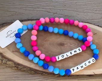 Puede incluir: Dos pulseras de cuentas con un degradado de colores de azul a rosa. Una pulsera deletrea "JOLENE" y la otra "PANCHO" con cuentas de letras blancas. Las pulseras están sobre una superficie de madera, con una etiqueta blanca "addykate".