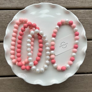 May include: Two necklaces on a white plate. One necklace is made of pink and light pink silicone beads. The other necklace is made of white pearl beads. The necklaces are on a white plate with a scalloped edge. A small white tag with the text "addykate" is on the plate.
