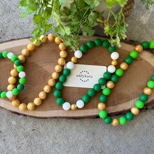 May include: Three silicone beaded bracelets in green, white, and gold. The bracelets are arranged on a wooden surface. The text "addykate" is visible on a white card.