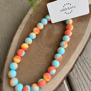 May include: A colorful necklace made of round beads with a blue, orange, and yellow swirl pattern. The necklace is on a wooden surface with a white card that says "addykate" on it.