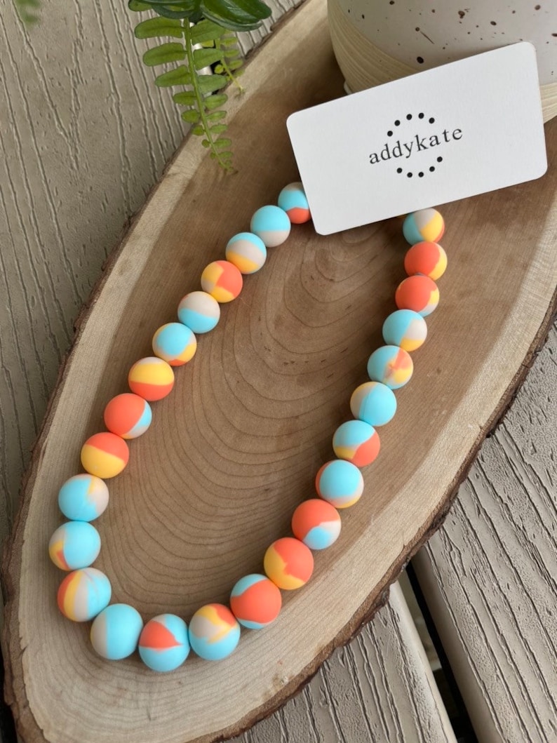 May include: A colorful necklace made of round beads with a blue, orange, and yellow swirl pattern. The necklace is on a wooden surface with a white card that says "addykate" on it.