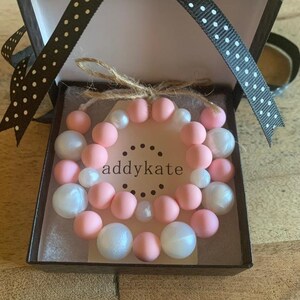 May include: A pink and white beaded bracelet set in a black gift box with white polka dots. The box is tied with a black ribbon and the bracelet is labeled "addykate".